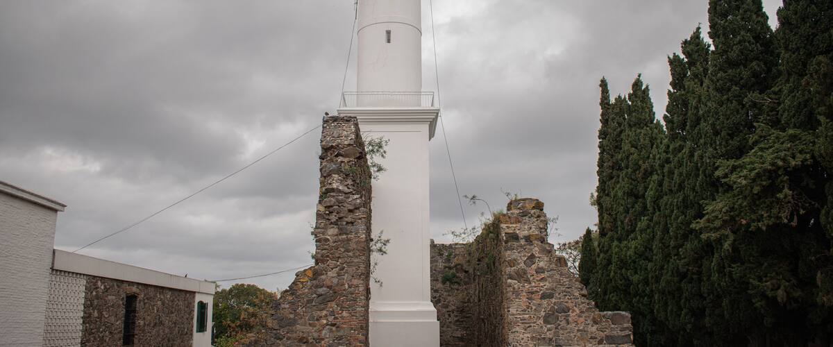 A white lighthouse with a stone base, surrounded by trees and a cloudy sky.