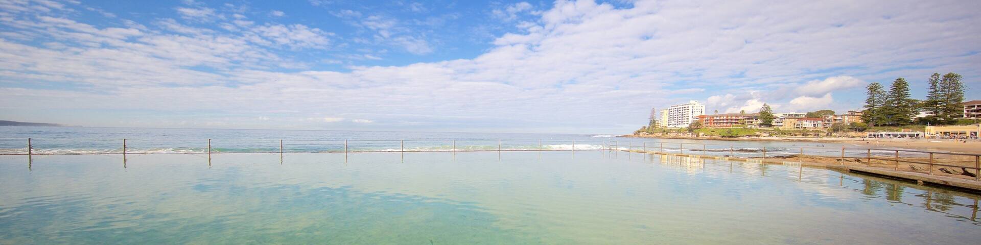 A clear pool with a wooden deck, surrounded by a beach and buildings.