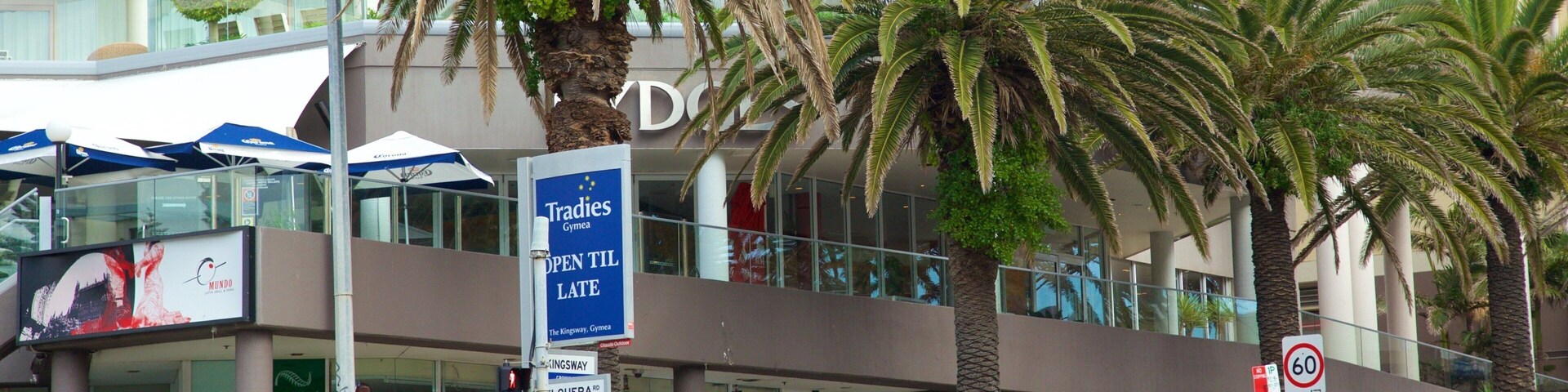 Un edificio moderno con palmeras frente a él. Hay un letrero que dice 'Tradies Gym' y una señal de límite de velocidad de '60'.