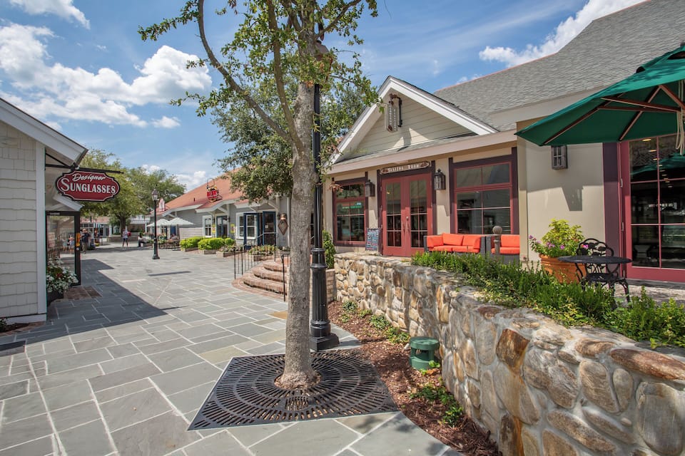 A stone-paved walkway with a tree, a stone wall, and a building with a sign for 'Designer Sunglasses'.