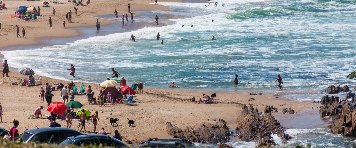A crowded beach with people swimming and sunbathing, umbrellas, and beachside buildings.