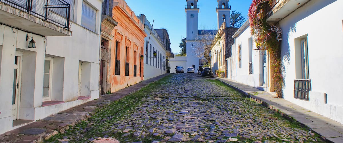 A cobblestone street flanked by historic buildings, a church with two towers, and a clear blue sky.