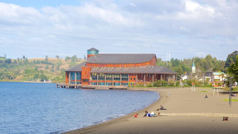 Un bâtiment situé au bord de l’eau, avec un quai, une plage où se trouvent des gens, et une église en arrière-plan.