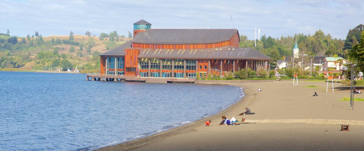 A waterfront building with a dock, a beach with people, and a church in the background.