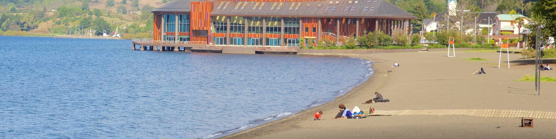 A waterfront building with a dock, a beach with people, and a church in the background.
