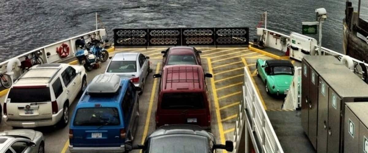 A ferry with cars parked on the deck, a body of water, and a cloudy sky in the background.