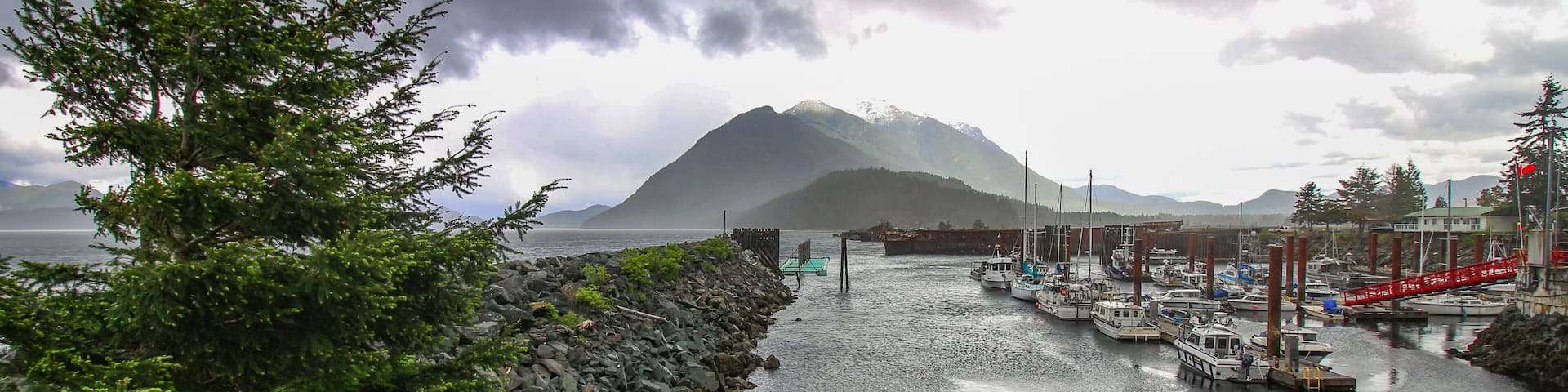 Kelsey Bay - north shore of Vancouver Island, BC. The view on the harbour, boats, rocks, pine tree, pier and mountains in the background. Stormy dark sky.