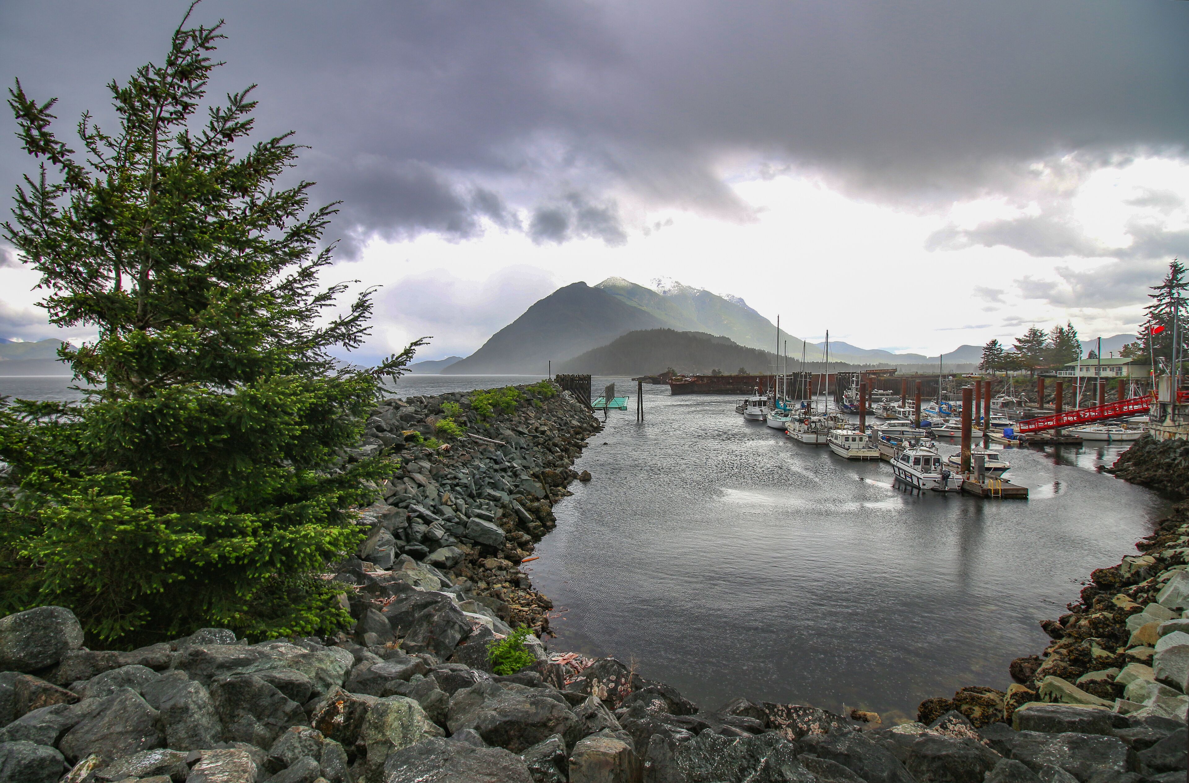 A rocky shoreline with a row of boats docked at a pier, a mountain in the background, and a cloudy sky.