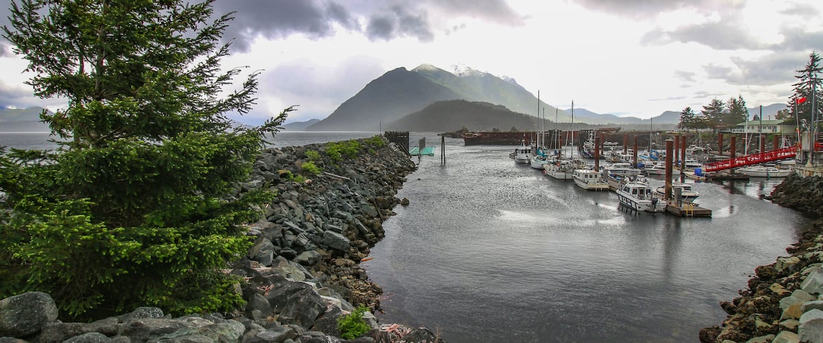 A rocky shoreline with a row of boats docked at a pier, a mountain in the background, and a cloudy sky.