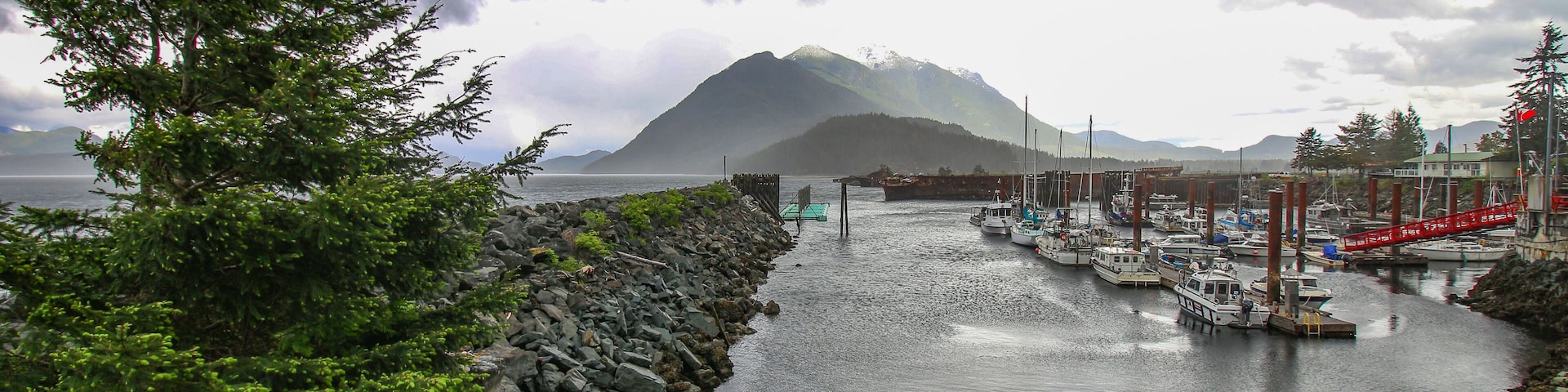 A rocky shoreline with a row of boats docked at a pier, a mountain in the background, and a cloudy sky.