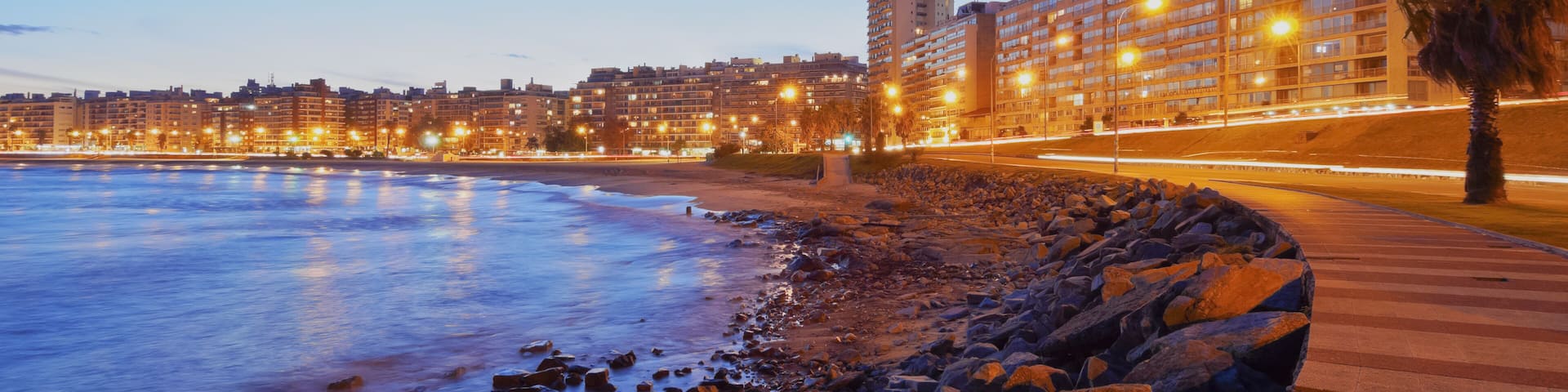 A coastal cityscape at dusk with illuminated buildings and a rocky shoreline.