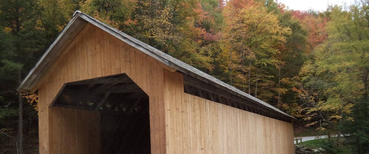 A wooden covered bridge with a wooden roof, surrounded by autumn-colored trees and a rocky stream.