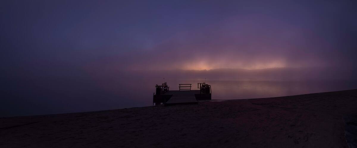 A beach with a wooden bench and chairs facing the water, under a dusky sky.