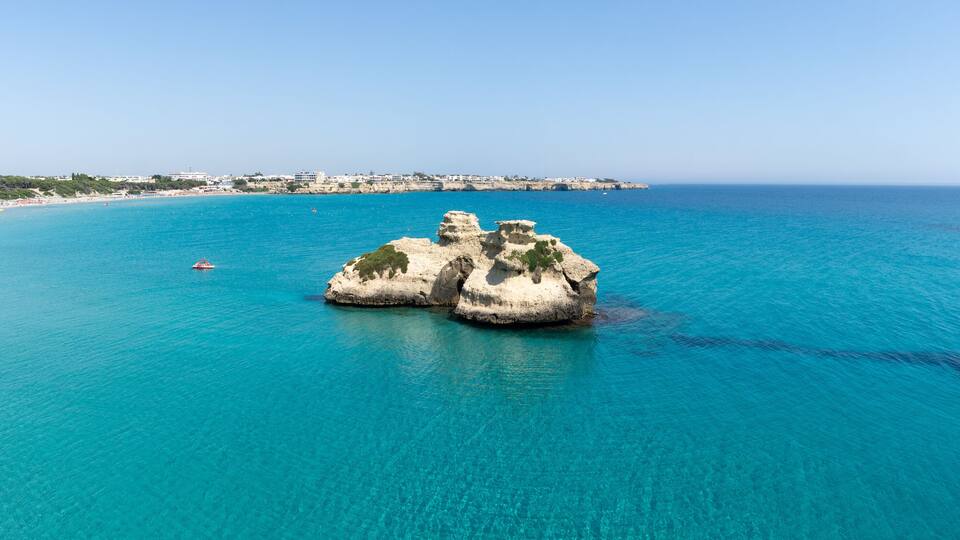 A coastal landscape with a prominent rock formation surrounded by turquoise waters. In the background, there is a beach with buildings and a clear sky.