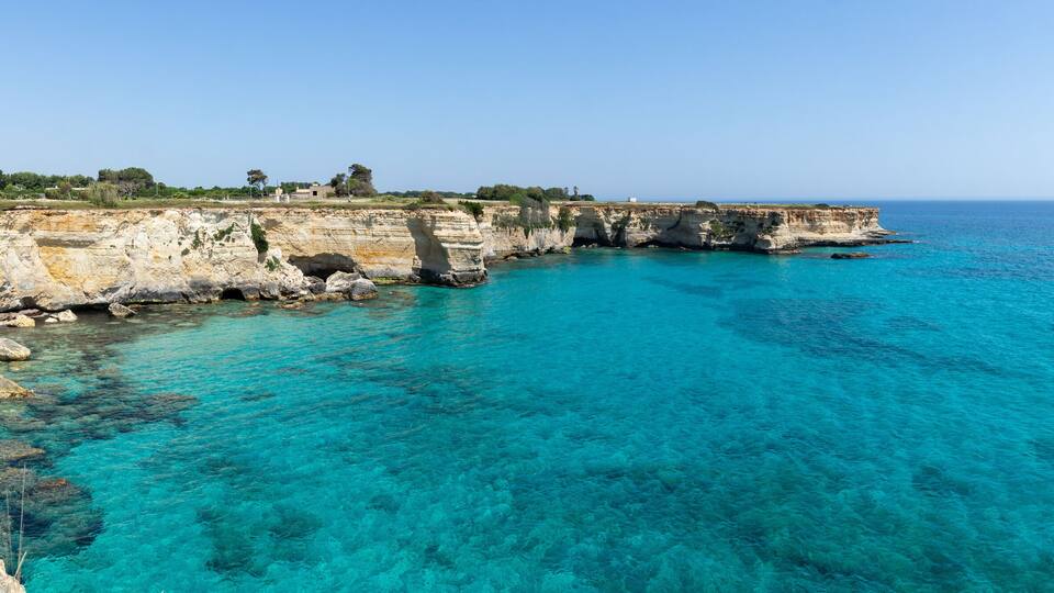 A coastal cliff with clear turquoise waters and a distant shoreline.