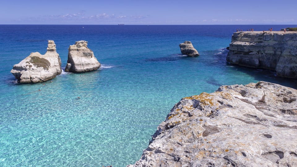 A coastal landscape with clear turquoise waters, rocky formations, and a distant ship on the horizon.