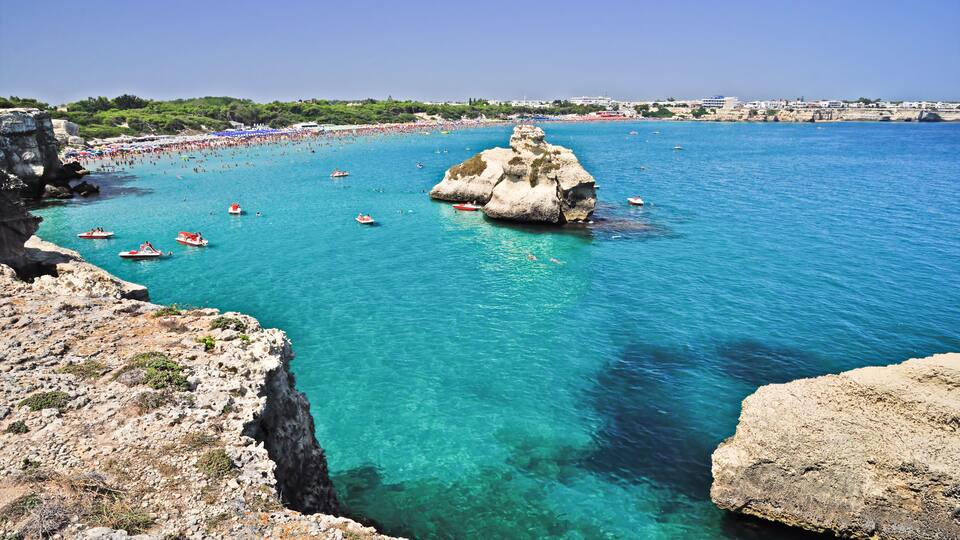 A coastal area with clear turquoise waters, rocky cliffs, and a large rock formation in the center.