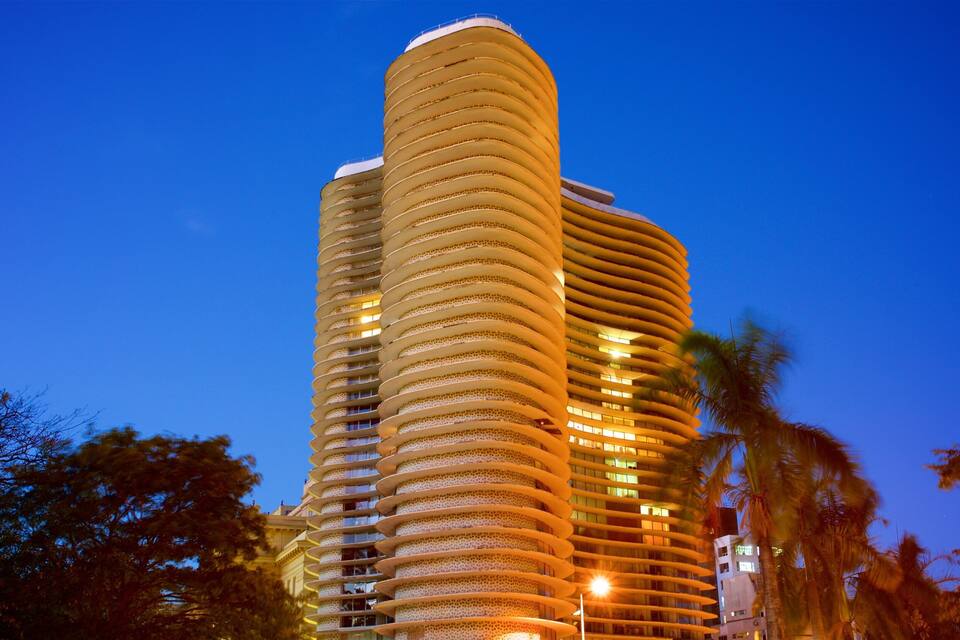 Un edifici alt i cilíndric amb un disseny en espiral, il·luminat durant la nit, envoltat de palmeres.