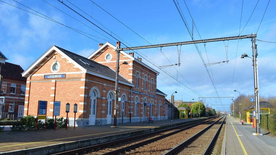 A train station with a brick building, tracks, overhead wires, and a clear sky.