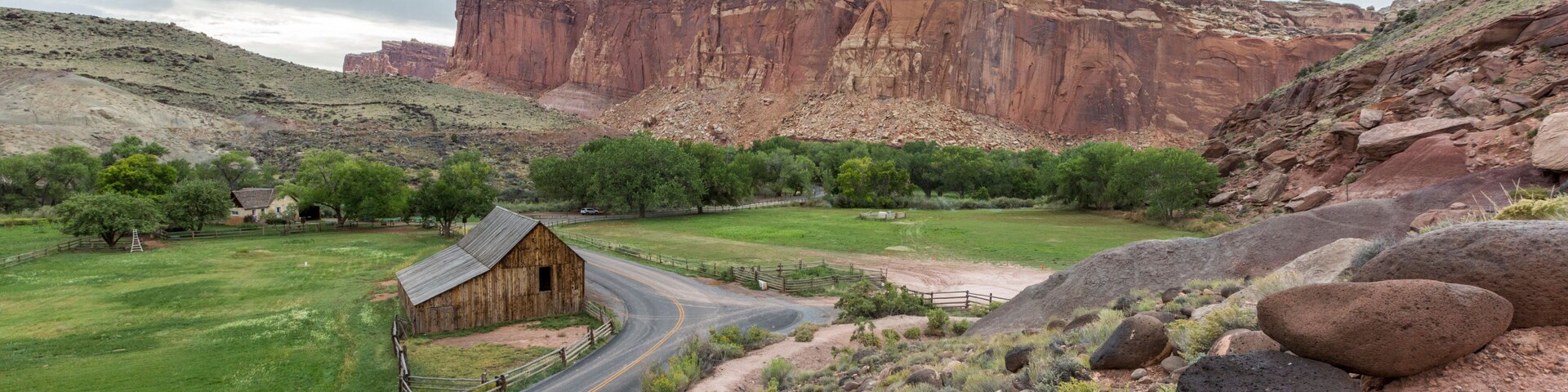 Paisaje rural con un camino sinuoso, un granero de madera y formaciones rocosas rojas al fondo.