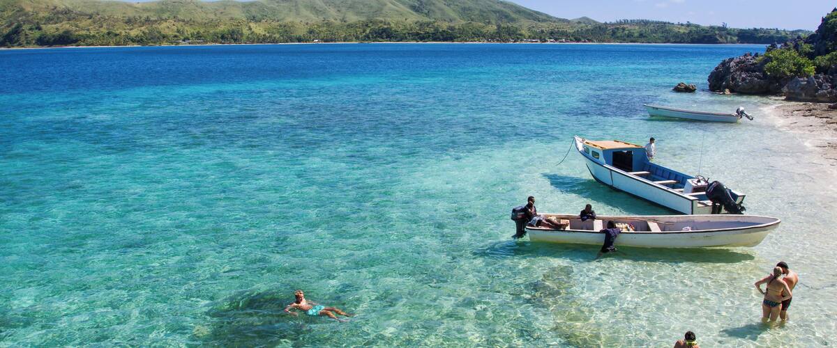 A tropical beach with clear turquoise water, boats, and people enjoying the water.