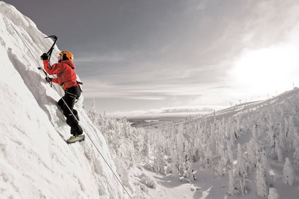 Une personne escalade une montagne enneigée, avec une vallée boisée en contrebas.