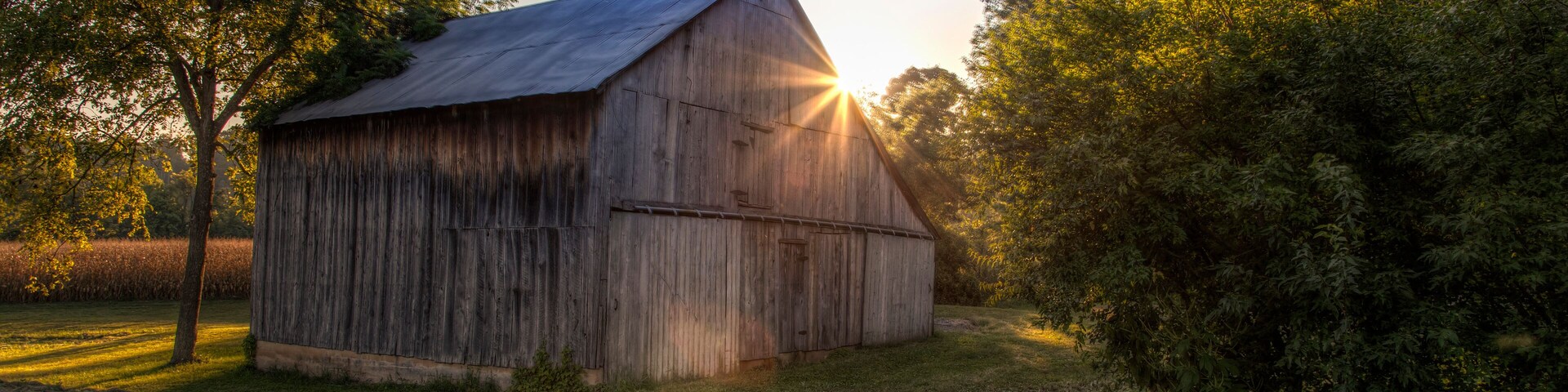 A wooden barn with a sloped roof, surrounded by trees and grass, with the sun setting in the background.