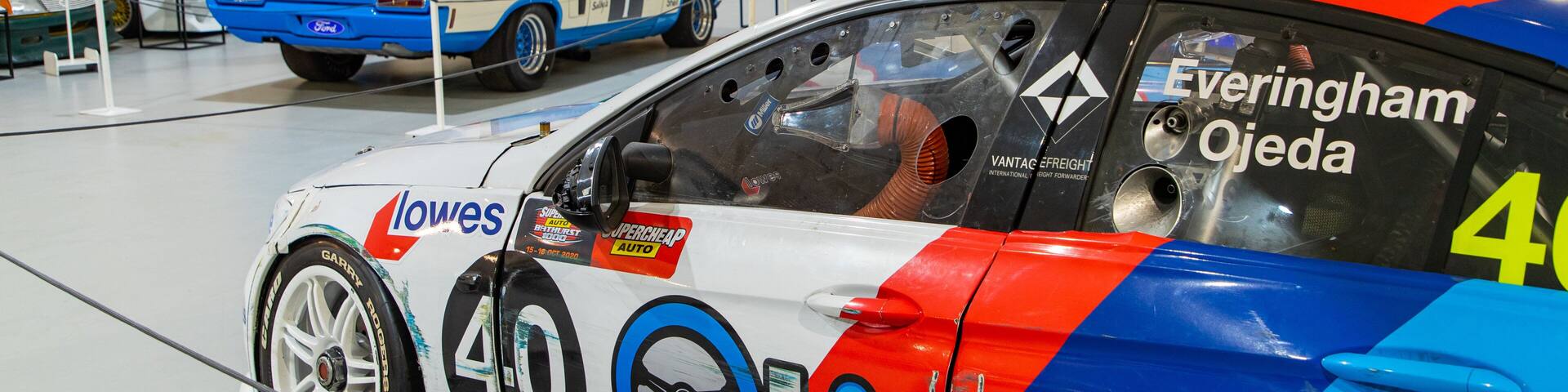 A racing car with the number 40, adorned with various sponsor logos, parked in a museum with other classic cars.