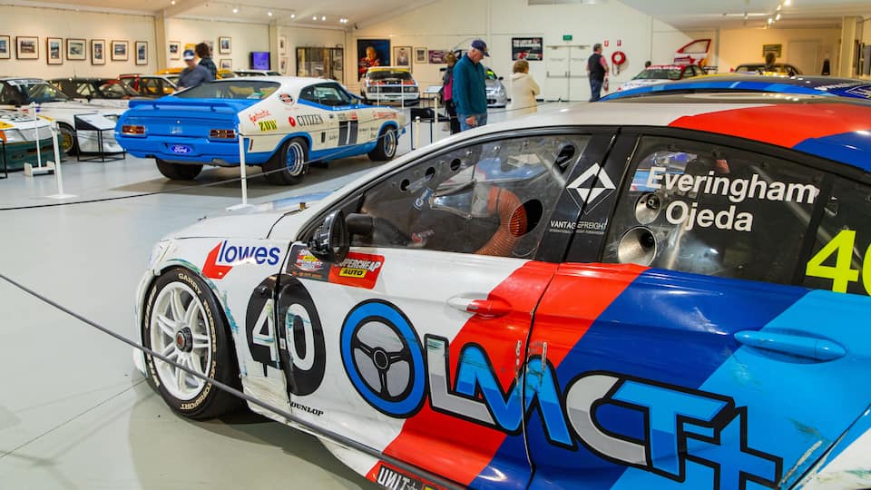 A racing car with the number 40, adorned with various sponsor logos, parked in a museum with other classic cars.
