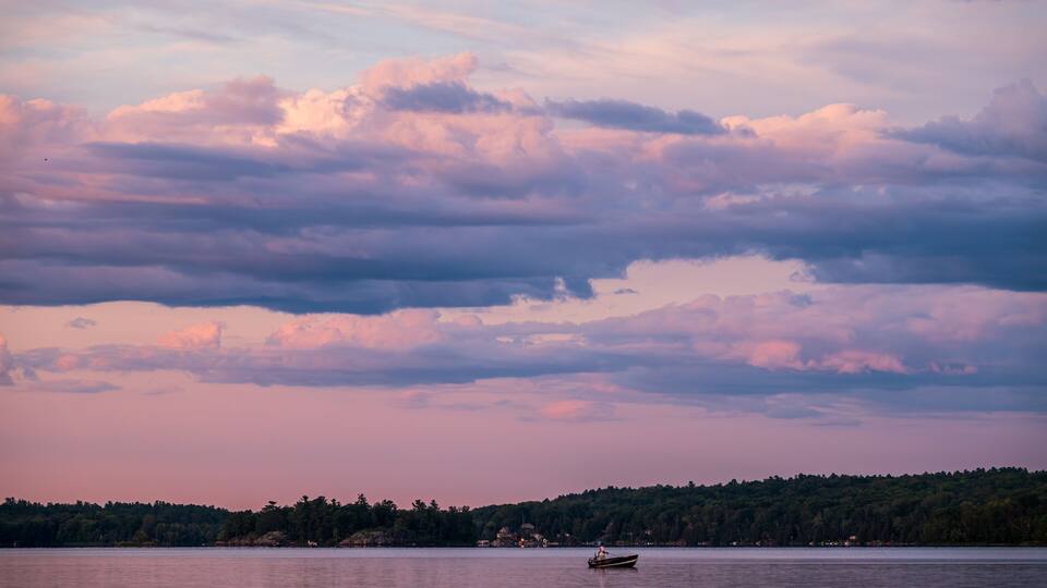 A calm lake with a boat in the center, surrounded by a forested shoreline and a cloudy sky.
