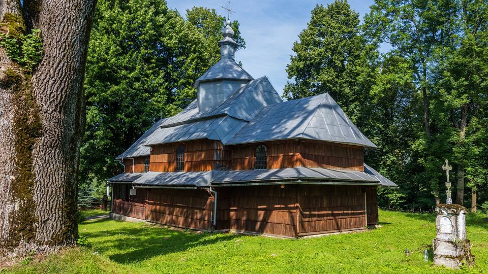A wooden church with a steeply pitched roof and a bell tower, surrounded by lush green trees.