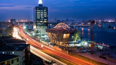 A cityscape at dusk with illuminated buildings, a prominent tower, and a bridge with light trails.