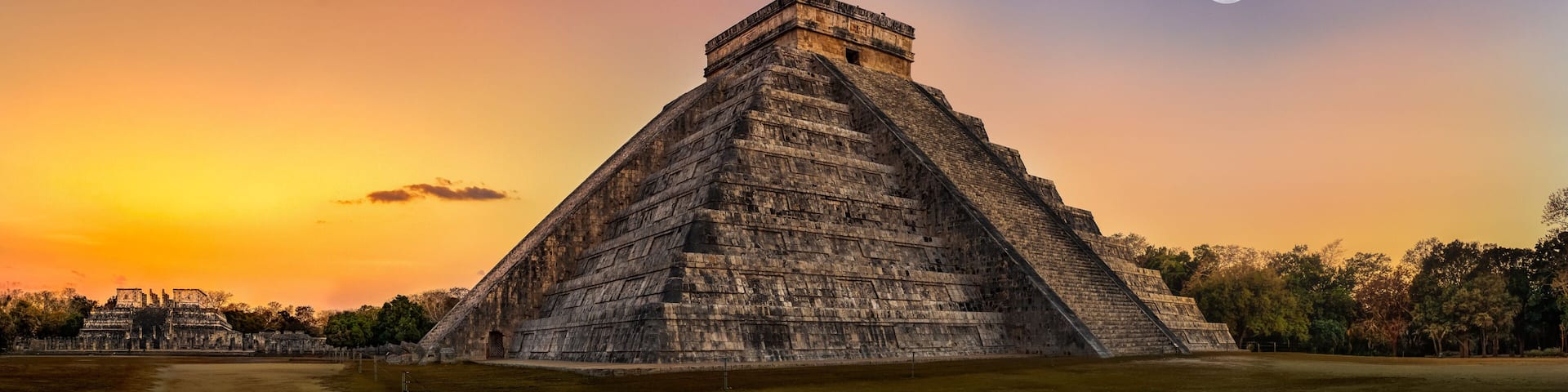 A large pyramid with a flat top, surrounded by a grassy area and trees, under a sunset sky with a visible moon.