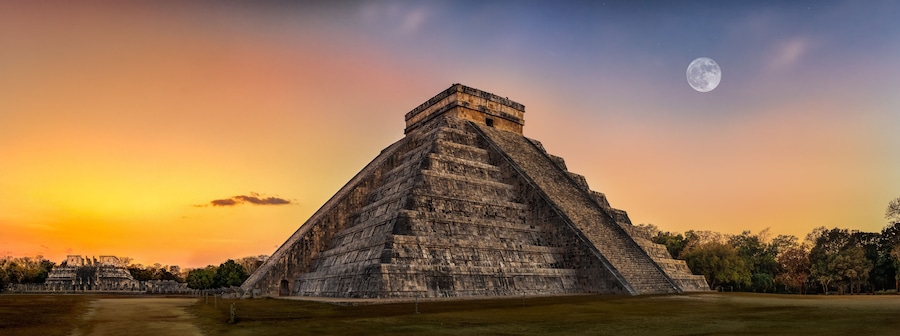 A large pyramid with a flat top, surrounded by a grassy area and trees, under a sunset sky with a visible moon.