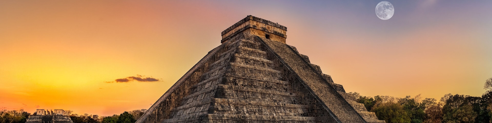 A large pyramid with a flat top, surrounded by a grassy area and trees, under a sunset sky with a visible moon.
