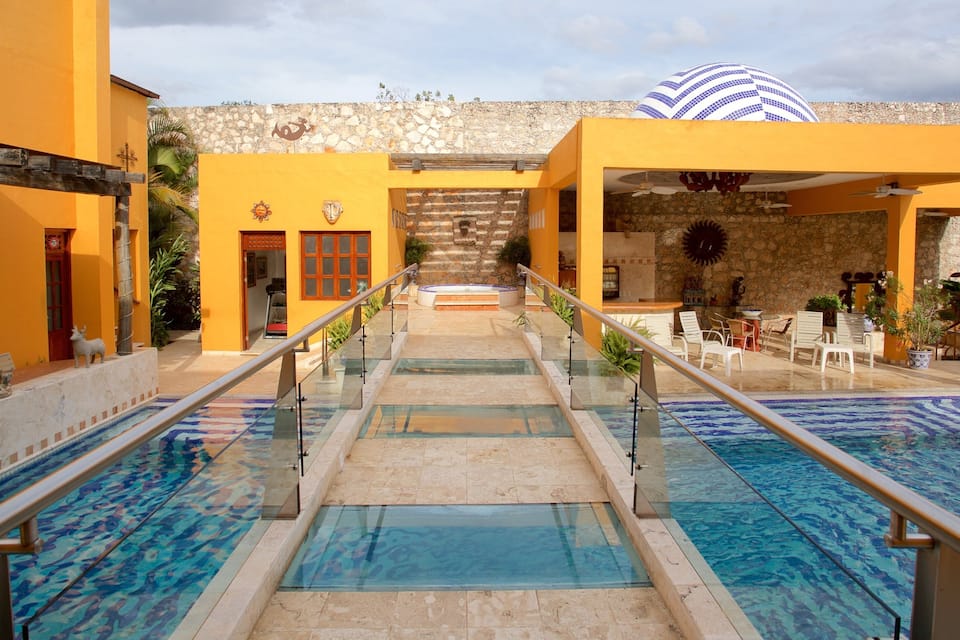 A swimming pool with a glass floor, surrounded by yellow buildings and a stone wall.