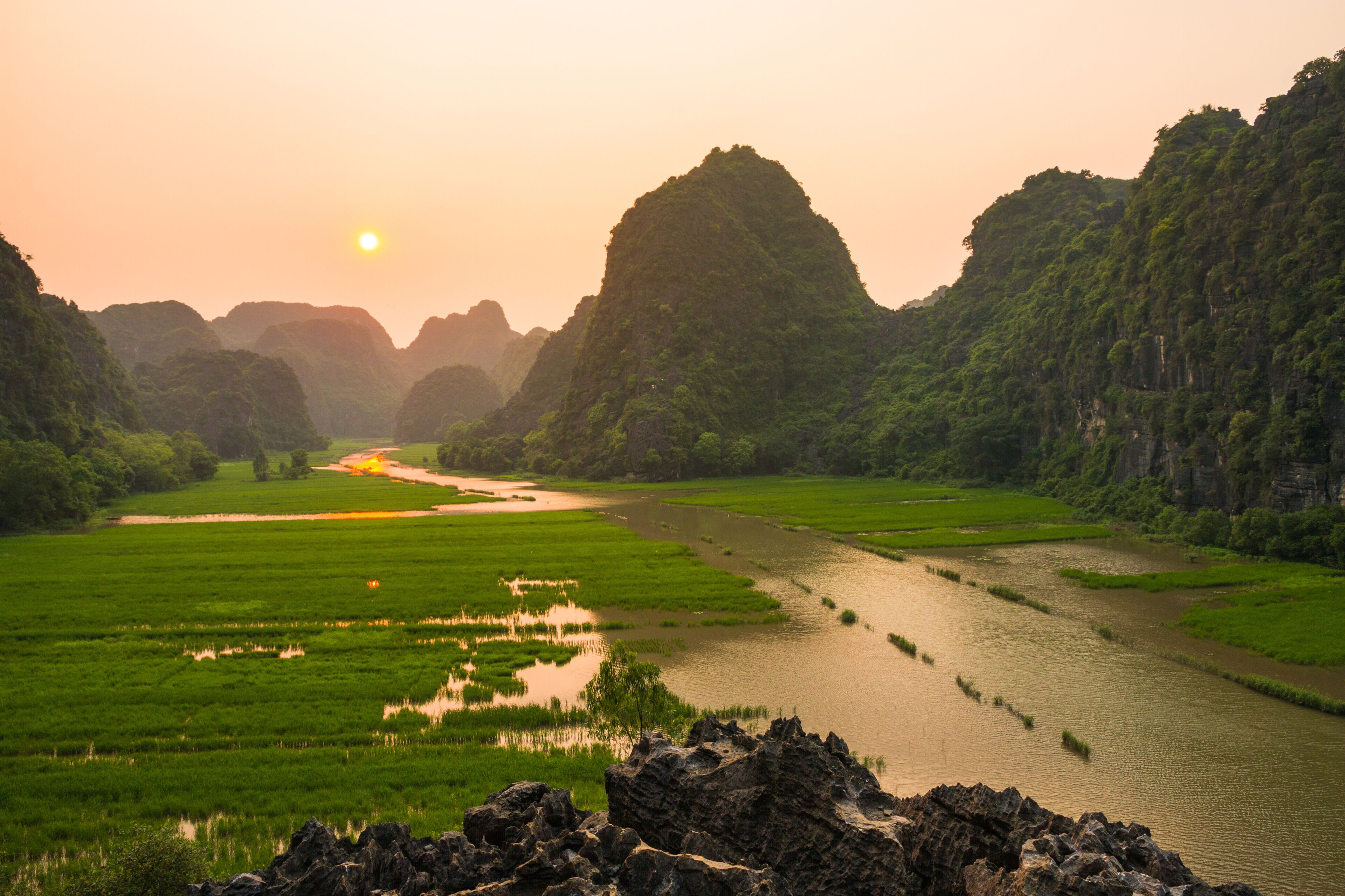 A river winding through lush green fields and limestone karsts under a setting sun.