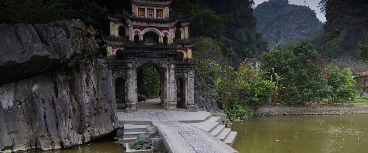 A traditional Chinese gate with a stone bridge leading to it, surrounded by lush greenery and a calm river.