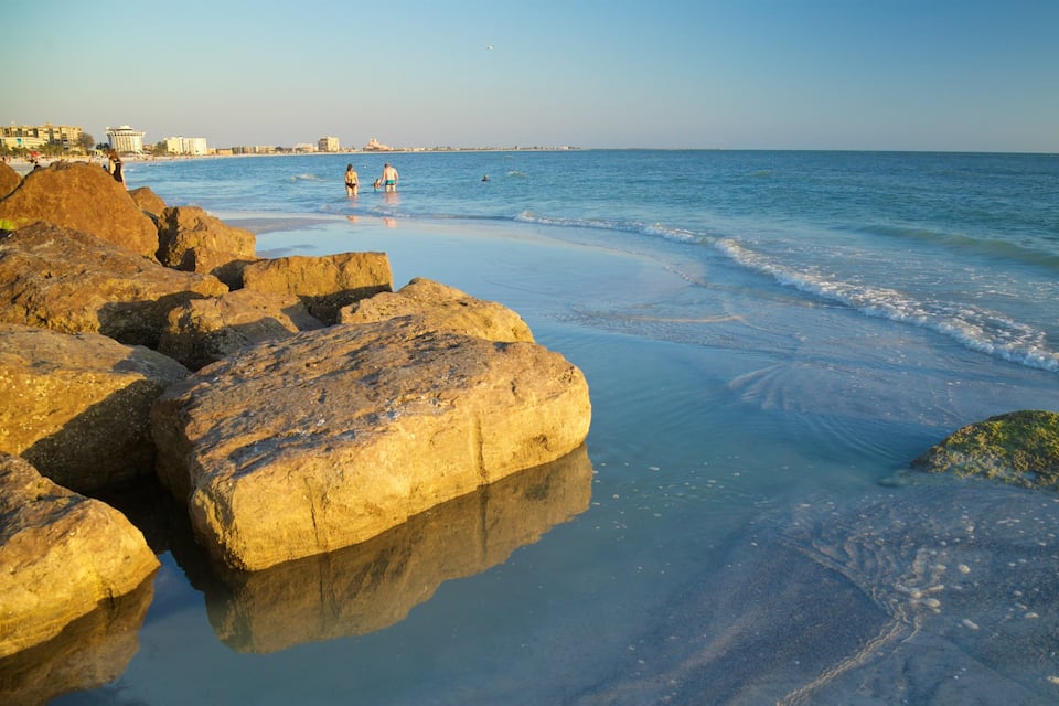 A beach with large rocks, calm water, and a distant shoreline with buildings.