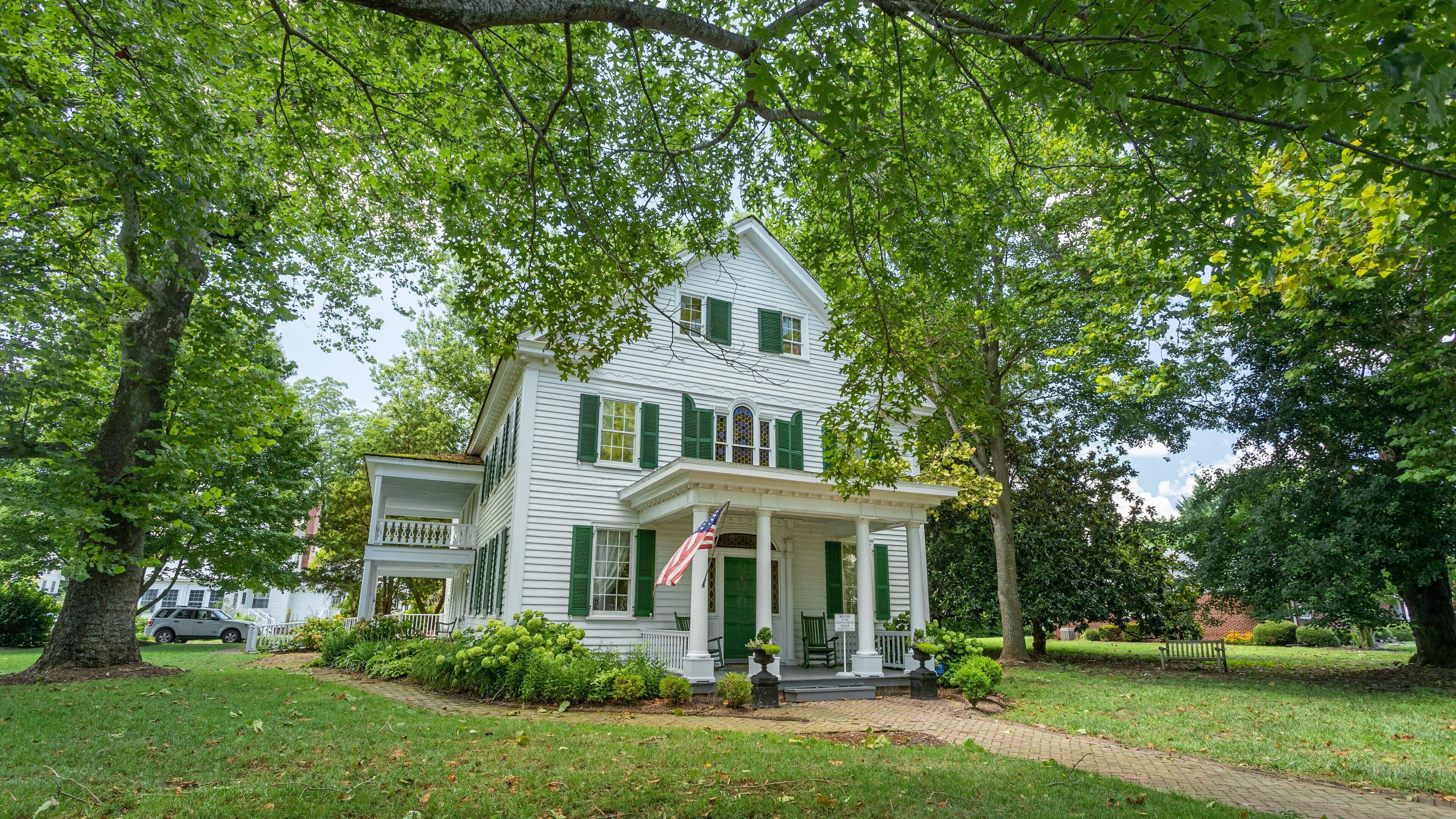 Une maison blanche de style colonial, dotée de volets verts, entourée d’une végétation luxuriante et bordée d’un chemin en briques.