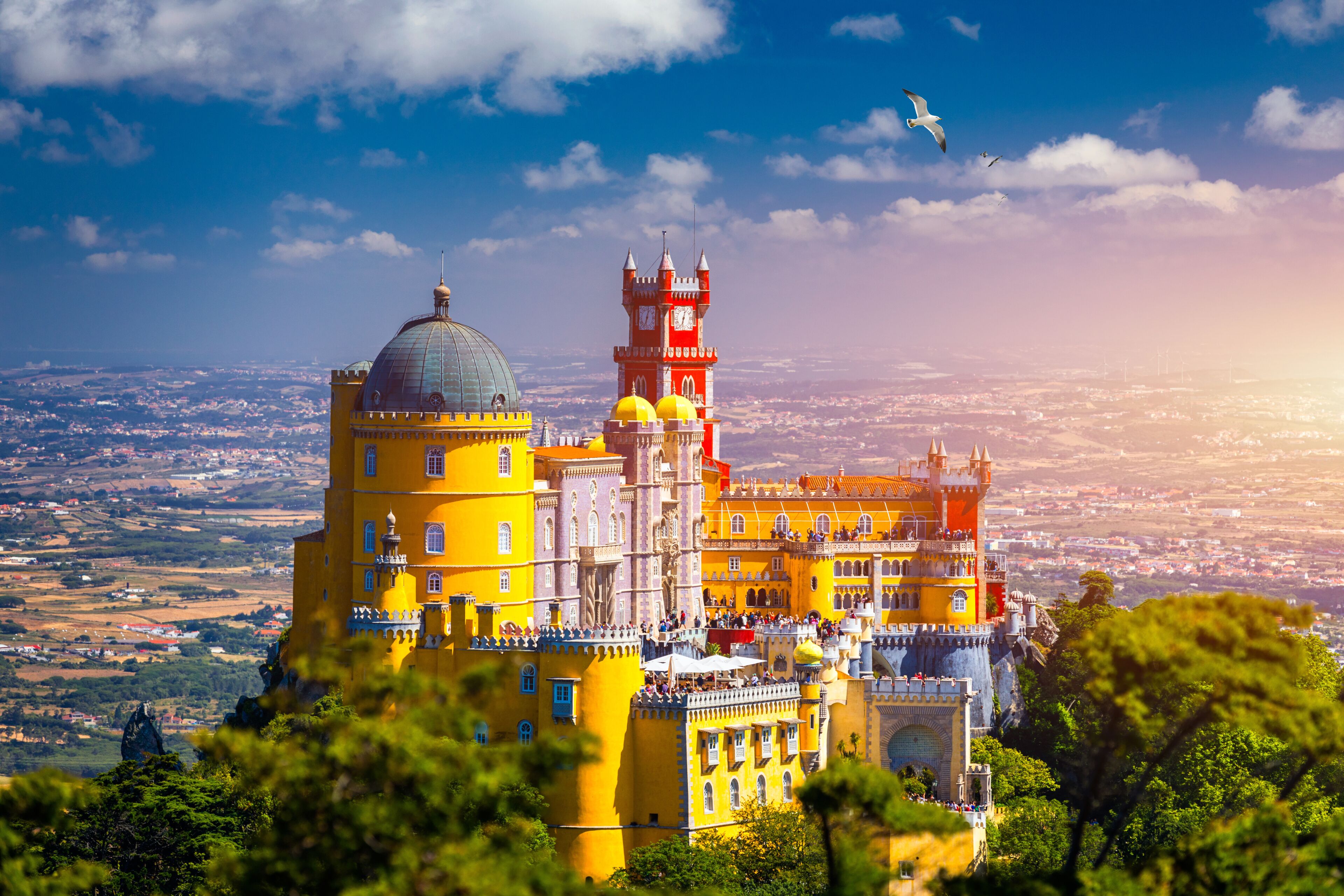 A colorful castle with a prominent tower and dome, surrounded by lush greenery and a panoramic view of the landscape.
