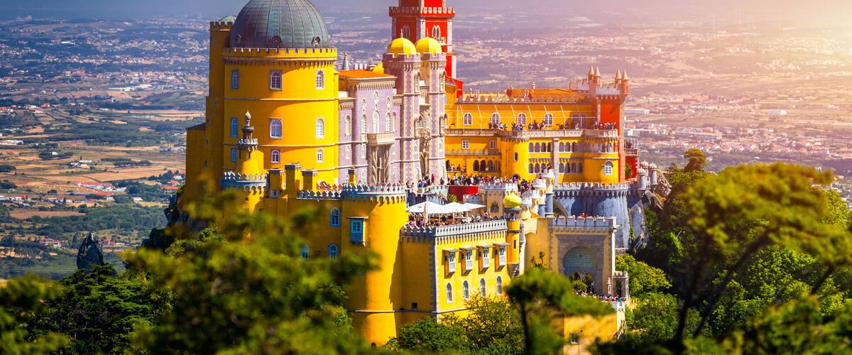 A colorful castle with a prominent tower and dome, surrounded by lush greenery and a panoramic view of the landscape.