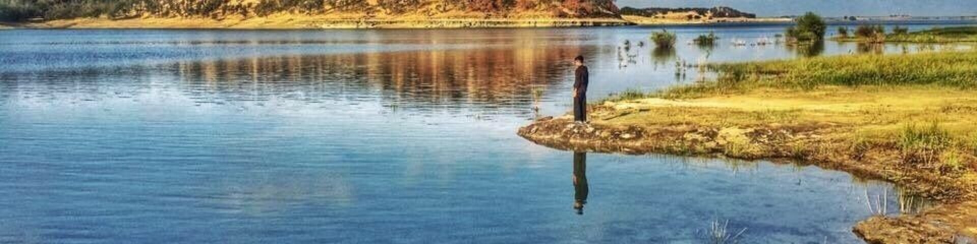 Un lago tranquilo con aguas cristalinas, una persona de pie en una pequeña isla y una colina con árboles al fondo.