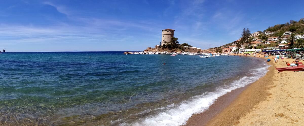 A beach with clear water, a tower, and houses on a hillside.
