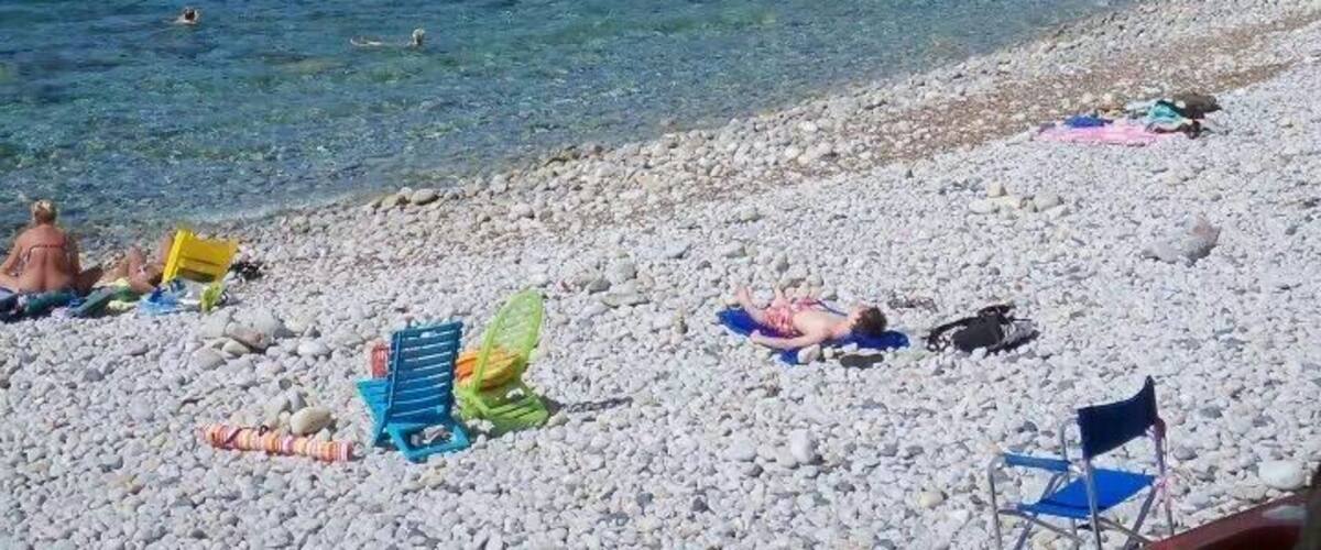 A pebble beach with clear blue water, people sunbathing, and a rocky hillside in the background.