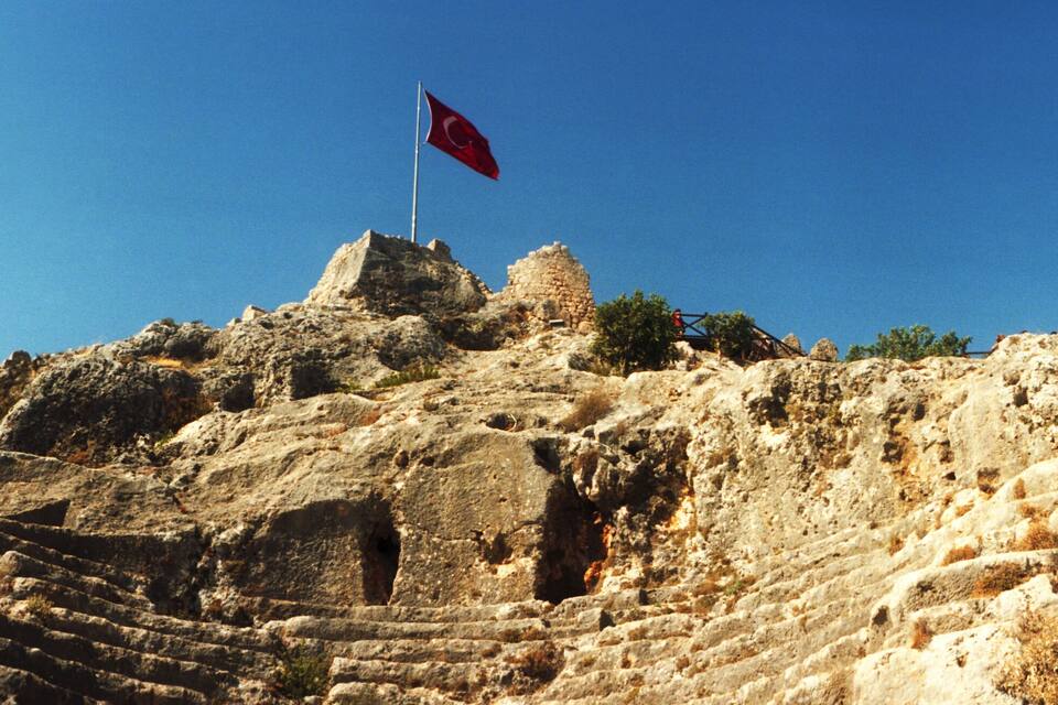 A rocky hill with a flag, stone ruins, and a wooden bridge.