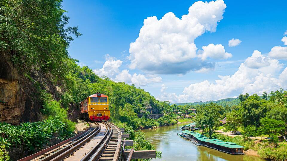 A yellow train on a curved railway track surrounded by lush greenery and a river.