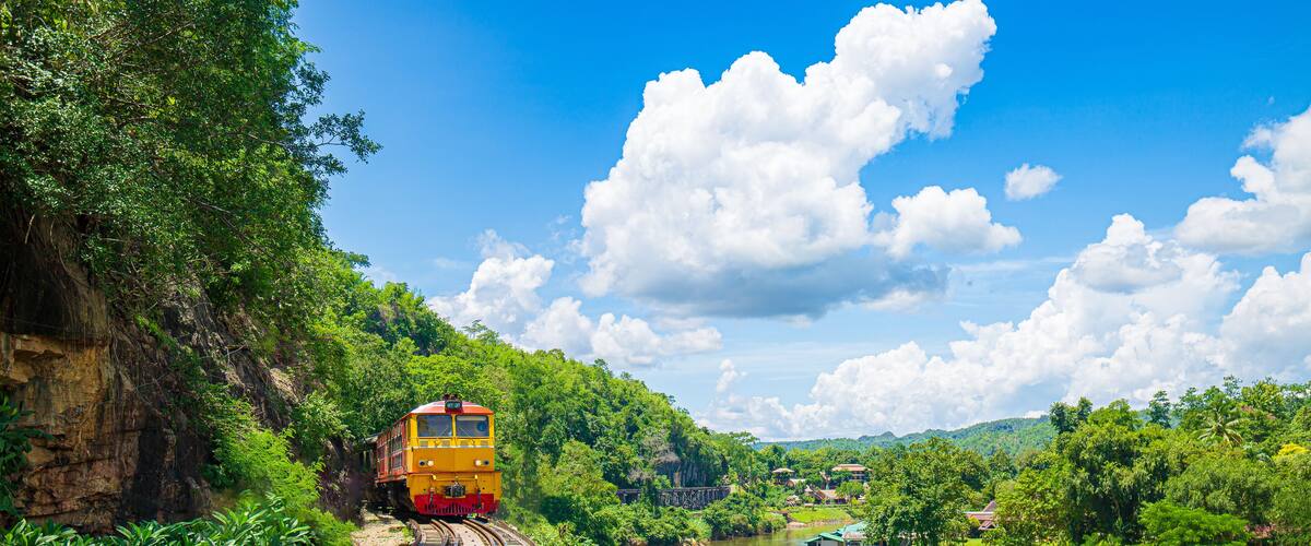 A yellow train on a curved railway track surrounded by lush greenery and a river.