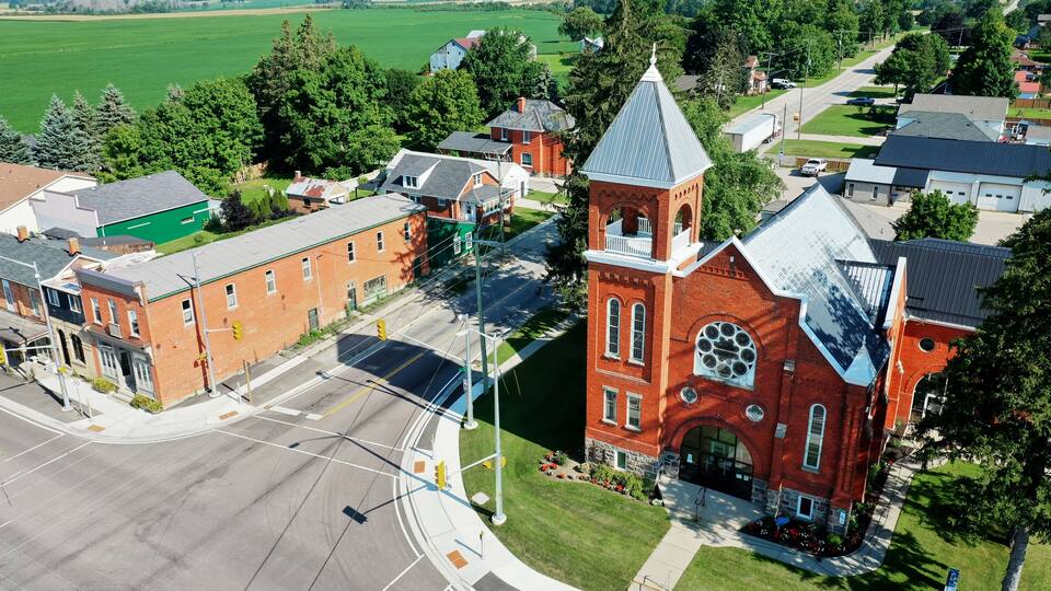 A red brick church with a steeple, surrounded by residential buildings and a green field.