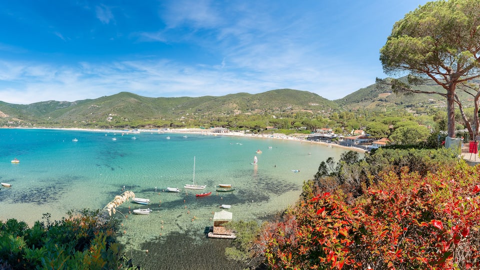 A coastal landscape with clear turquoise waters, boats, and a backdrop of green hills.